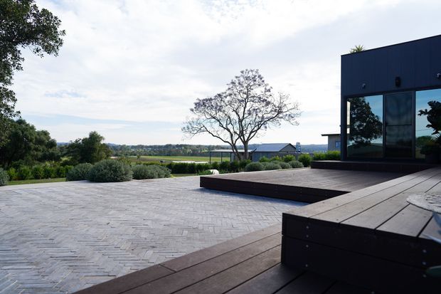 A contemporary dark-clad building opens onto a broad paved terrace and timber deck overlooking a landscaped rural or semi-rural setting. The outdoor space includes raised deck platforms, low planting, large windows, and a mature sculptural tree, creating a spacious connection between architecture and landscape.