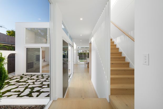 Bright contemporary residential hallway with pale timber flooring, white walls, recessed ceiling lights, and a straight timber staircase enclosed by vertical white slatted screening. Full-height glazing opens to a landscaped courtyard with irregular stone paving and greenery, while a dining or living area is visible at the far end.