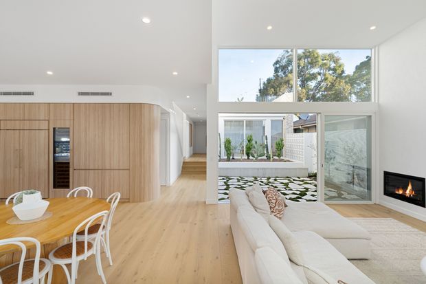 Bright open-plan contemporary interior combining a dining area, kitchen cabinetry and living room. The space features pale timber flooring and curved timber kitchen joinery, white walls and ceiling, expansive glazing to a planted courtyard, a light sectional sofa, and a recessed linear fireplace.