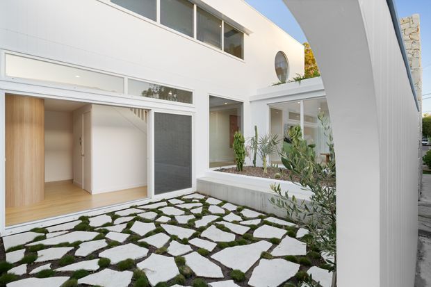 Bright contemporary two-storey courtyard exterior with crisp white rendered walls, extensive glazing, a circular window, and a covered outdoor passage. Irregular pale stone pavers set in moss create a naturalistic courtyard surface, with raised planted beds and glimpses of light timber flooring and interior spaces through open glazing.