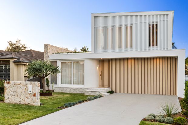 A contemporary two-storey suburban house with a white rendered facade, large vertically screened upper-level glazing, a broad timber-toned garage door, stone-clad lower walls and a pale concrete driveway. The landscaped front yard includes low ornamental planting and a stone mailbox feature.