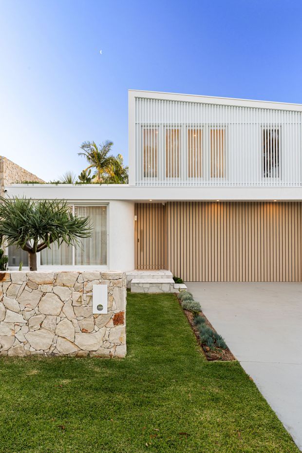 Front exterior view of a contemporary two-storey residence with a crisp white facade, upper-level vertical slatted screening, warm timber-toned garage and entry cladding, pale stone boundary wall, landscaped lawn, and a concrete driveway. Tropical planting and clear blue sky frame the modern street elevation.