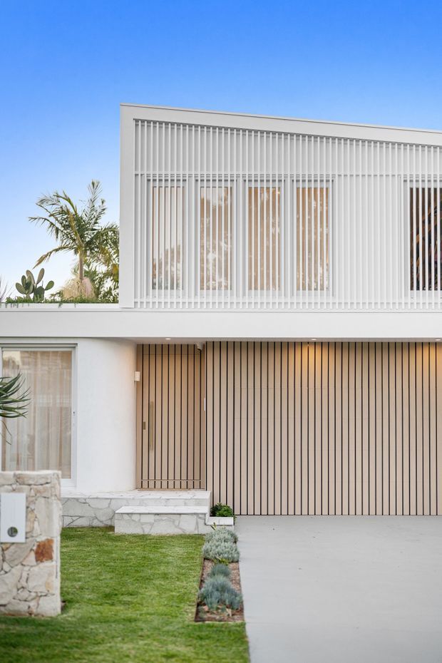 Exterior view of a contemporary two-storey residence with a clean white rendered facade, extensive vertical white screening over the upper-level glazing, and a matching vertically slatted garage door and entry door. Pale stone retaining and entry walls, a concrete driveway, manicured lawn, and drought-tolerant planting complete the frontage.