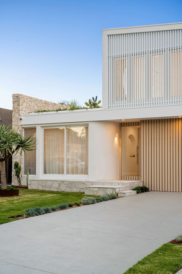 Street-facing view of a refined modern two-storey residence with a white rendered facade, large upper-level vertical slatted screening, pale stone feature walls and base cladding, timber-toned entry and garage screening, and a broad concrete driveway. Low, drought-tolerant landscaping and a manicured lawn frame the frontage.