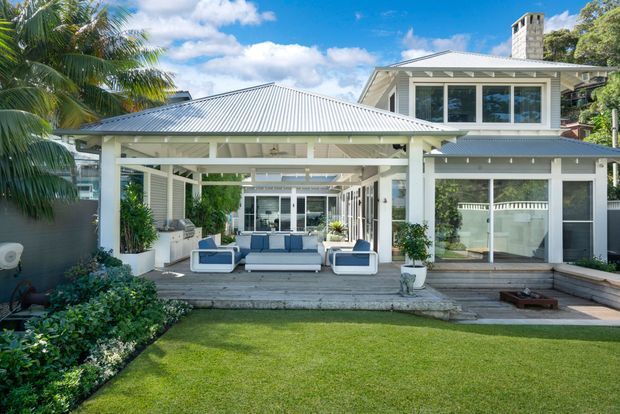 Exterior view of a bright, multi-level coastal-style home opening onto a covered outdoor living area. The house features white weatherboard cladding, extensive white-framed glazing, a light corrugated metal roof, timber decking, and a landscaped lawn with tropical planting. Outdoor sofas and a dining area occupy the covered patio.
