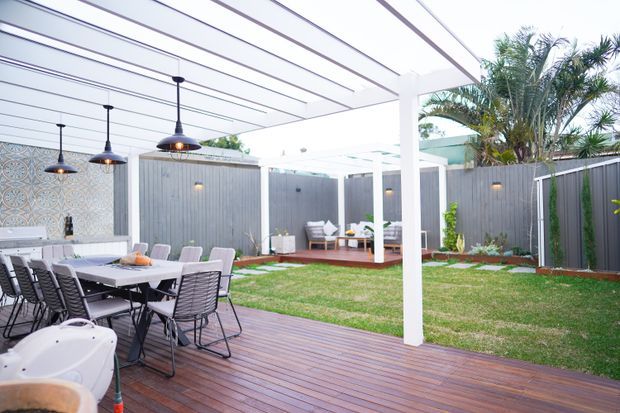 A bright outdoor entertaining area with a large timber deck, dining table and chairs, a covered pergola with linear roof battens, pendant lights, and a separate timber lounge platform. Grey vertical fencing, patterned feature tiling, lawn, and tropical planting create a contemporary backyard setting.