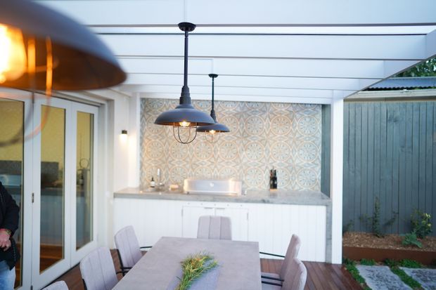Covered outdoor dining and entertaining area with a long dining table, upholstered chairs, pendant lighting, and a built-in outdoor kitchenette or bar. The space features a decorative patterned tile backsplash, pale cabinetry and benchtop, white-framed glazed doors, timber decking, and a vertical timber privacy screen with landscaped planting.