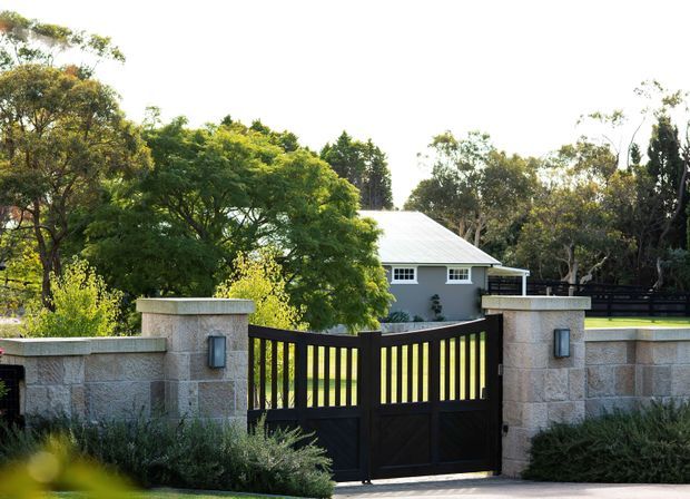 A landscaped residential property viewed from the street, with a dark timber driveway gate set between pale textured masonry pillars and low walls. Behind the gate is a single-storey light-coloured house with a low pitched metal roof, surrounded by mature trees and garden planting.