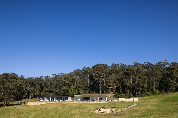 A low, elongated contemporary single-storey residence set within a broad grassy landscape at the edge of dense native woodland. The home has dark glazed frontage, pale rendered walls, and a simple low-pitched roofline. A curved stone-edged garden feature leads through the foreground, beneath a clear blue sky.