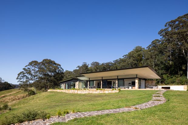 A low, single-storey contemporary residence set into a gently sloping grassy site, with a broad flat overhanging roof, extensive glazing, pale stone walls, and a paved stepping path leading through the lawn. Dense native-looking trees form a backdrop.