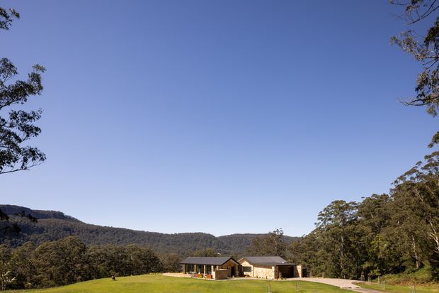 A low, single-storey rural residence set in an expansive green lawn, surrounded by dense native bush and rolling forested hills. The home has a simple contemporary form with dark pitched roofing, pale masonry walls, and a long, understated profile. A gravel or pale paved driveway leads toward the house, with a broad clear blue sky dominating the composition.