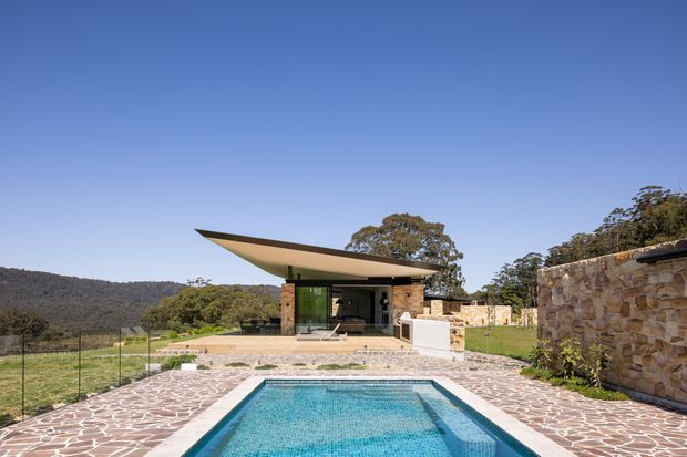 Contemporary single-storey pavilion set within a rural, wooded landscape. The building features a dramatic asymmetrical projecting roof, extensive glazing, stone walls, and a paved terrace surrounding a rectangular swimming pool. A low stone boundary wall frames the right side of the scene.