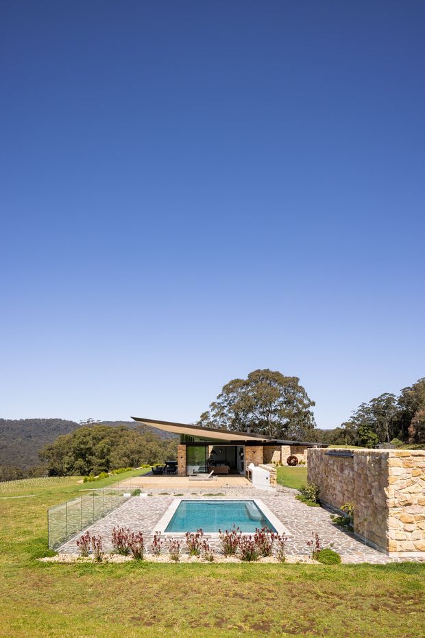A landscaped rural property viewed from the garden, featuring a rectangular swimming pool with glass balustrading and pale stone paving in the foreground. Beyond the pool is a low, contemporary pavilion with broad projecting roof planes, extensive glazing, and stone-clad walls. A substantial dry-stone boundary wall, lawn, mature trees, and wooded hills complete the scene under a clear blue sky.