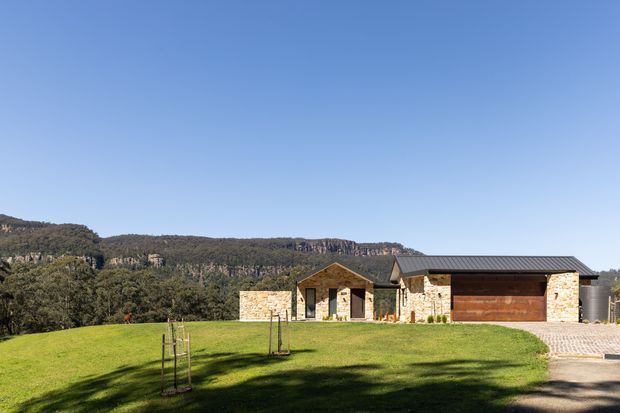 A low, single-storey contemporary residence set in a landscaped lawn, with pale stone walls, dark pitched roof sections, timber-clad entry and garage doors, and a paved driveway. A dramatic wooded escarpment forms the distant backdrop.