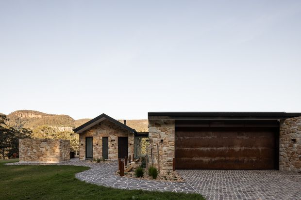 Exterior view of a low, stone-clad residential property in a rugged hillside setting. The composition features a detached gabled stone structure with narrow dark-framed windows, a larger flat-roofed stone garage with a broad dark timber or metal door, and a patterned stone-paved driveway and walkway. The architecture has a restrained contemporary rural character, with warm natural masonry integrated into the landscape.