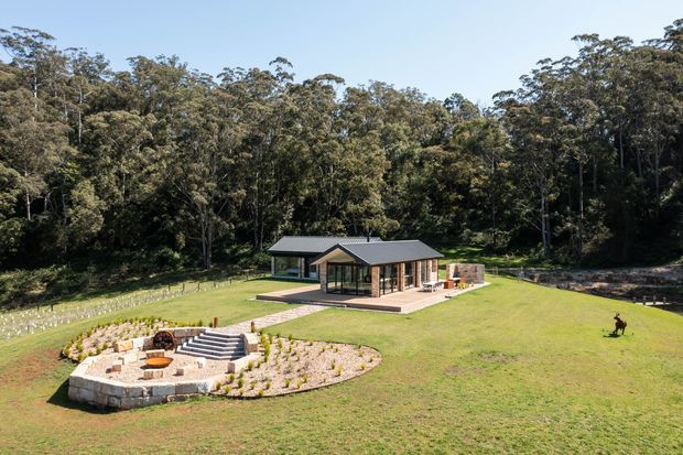 A low, modern single-storey pavilion-style home set within a broad landscaped lawn and surrounded by dense eucalyptus woodland. The building has dark metal pitched roofs, extensive floor-to-ceiling glazing, timber-toned structural elements, and a paved terrace. In the foreground is a circular outdoor fire-pit seating area with stone retaining walls and planted garden beds.