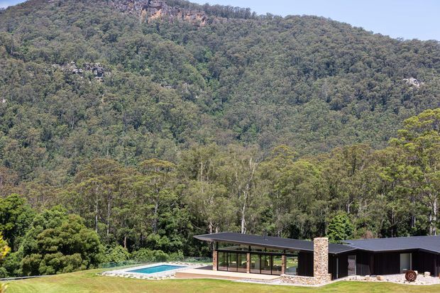 A low, modern single-storey house with dark flat roofing and extensive glazing sits at the edge of a landscaped lawn, with a long swimming pool in front. A dramatic densely forested mountain or escarpment rises directly behind the property.