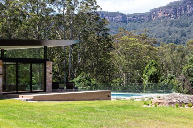A modern pavilion-style house set within a lush bushland landscape, with extensive dark-framed glazing, a thin projecting flat roof, timber-clad walls, and a swimming pool overlooking dense forest and dramatic sandstone cliffs.