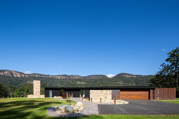 A low, single-storey contemporary residence set against a dramatic forested escarpment. The facade combines dark vertical cladding, warm stone masonry, extensive glazing, and a broad timber garage door, with a flat roofline and a paved driveway leading to the entry.