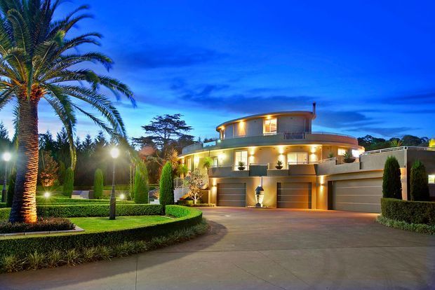 A large, highly landscaped luxury residence photographed at dusk. The home has a curved, multi-level form with extensive warm exterior lighting, broad glazing, balconies, and multiple integrated garage doors. Formal hedges, clipped columnar evergreens, a large palm, and a wide paved driveway frame the facade.