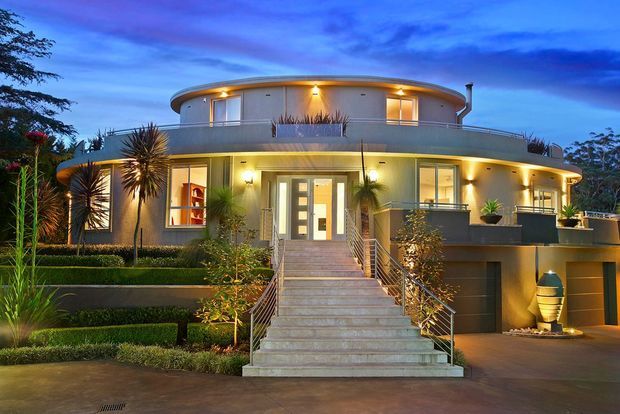 Exterior dusk photograph of a large, highly finished contemporary two-storey residence. The building has a rounded upper-storey form, curved balcony and roofline, pale rendered walls, extensive glazing, warm integrated exterior lighting, a broad central staircase with metal railings, landscaped terraces, and an integrated garage.