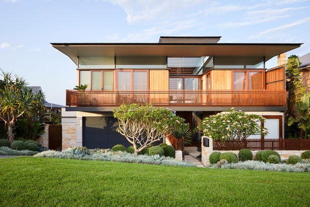 Front exterior view of a contemporary two-storey residence with a broad low-pitched roof, projecting upper-level balcony, timber-toned vertical screening and balustrades, extensive glazing, and a landscaped lawn with structured planting. The facade combines pale rendered walls, stone accents, dark garage doors, and warm timber elements.
