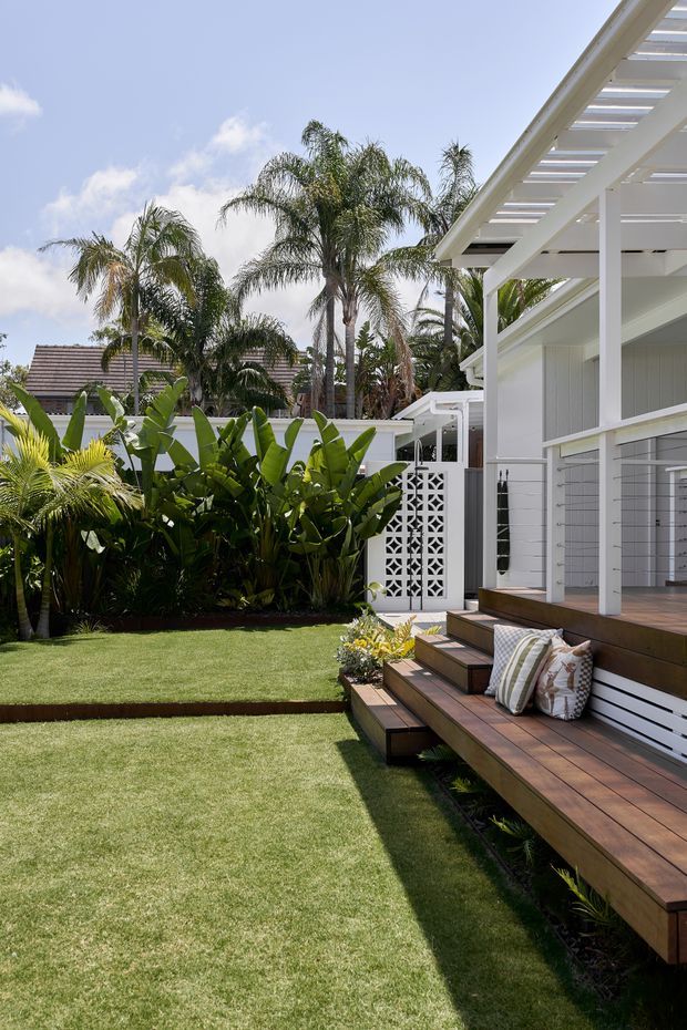 A bright landscaped backyard beside a white-clad house, featuring a raised timber deck with broad built-in bench seating, steps, and striped cushions. A manicured lawn, tropical palms and lush foliage surround a white decorative breeze-block privacy screen and covered outdoor structure.