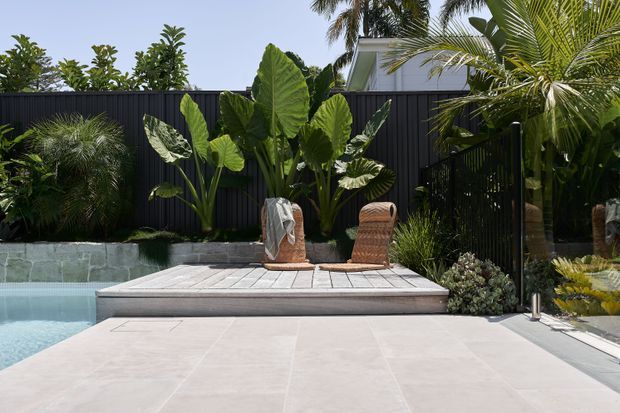 A contemporary outdoor pool area with a pale tiled terrace, raised timber deck, and two woven lounge chairs. Lush tropical planting, a dark vertical privacy fence, and a modern residential structure form the backdrop.