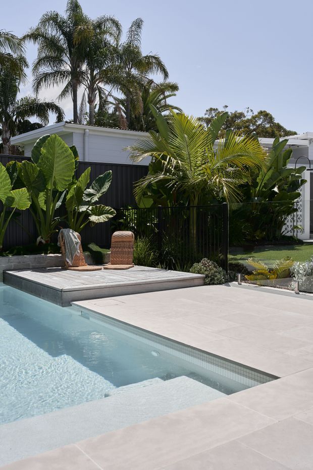 A bright contemporary backyard pool area with pale tiled paving, a light blue swimming pool, raised timber poolside platform, tropical planting, and a black privacy fence. A white low-profile building and decorative screen are visible behind the lush landscaping.