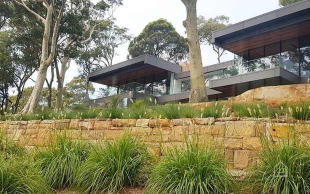 Exterior view of a contemporary multi-level residence integrated into a landscaped, sloping site. The home features strong horizontal flat roof planes, extensive glazing, dark metal framing, warm timber soffits, glass balustrades, and stepped natural-stone retaining walls surrounded by established trees and ornamental grasses.