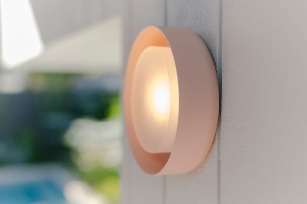 Close-up of a rounded wall-mounted exterior light fixture with a warm glowing bulb and peach-toned housing, attached to a pale vertically grooved wall. The background is softly blurred, with hints of greenery and an outdoor setting.
