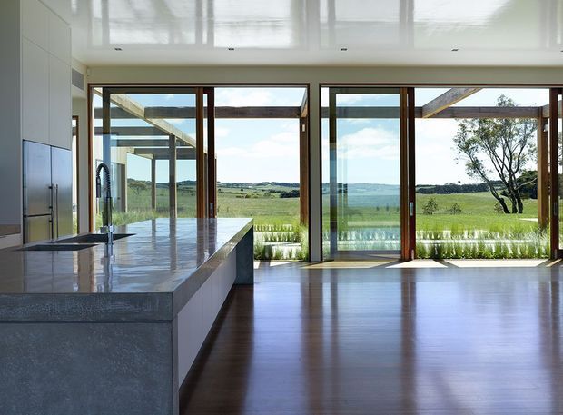 A bright, open-plan modern kitchen and living space with a large stone island, integrated cabinetry, polished white ceiling, and expansive timber-framed glazing. The glazing opens to a rural green landscape, while dark timber flooring and exposed outdoor structural beams add warmth and architectural interest.