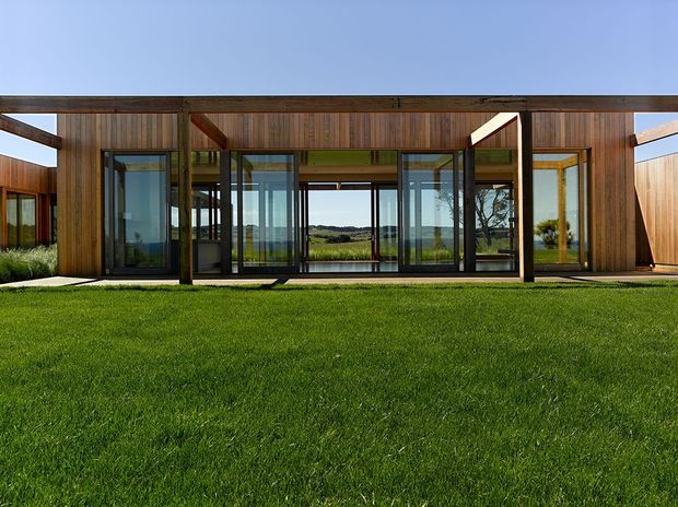 Single-storey contemporary residential pavilion viewed from a broad lawn, with extensive floor-to-ceiling glazing, dark window frames, and warm vertical timber cladding. The low horizontal form features sheltered recessed openings and a flat roofline, with open rural landscape visible through the glass.