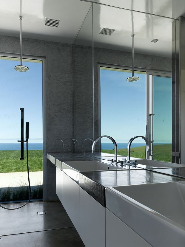 Contemporary bathroom with a freestanding bathtub, double vanity, expansive mirrors, large windows, and twin ceiling-mounted rain showers. The space has dark textured wall surfaces, glossy white built-in cabinetry, and a polished dark stone vanity top. Ocean and lawn views are visible through the glazing.