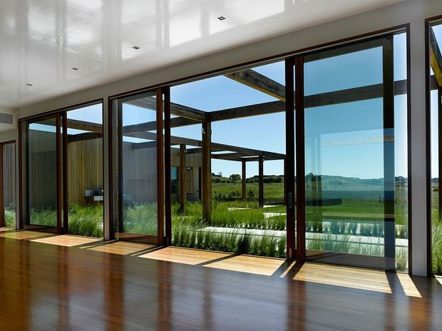 A bright contemporary interior with a polished dark timber floor, glossy white ceiling, and extensive floor-to-ceiling glazed sliding doors in dark timber frames. The glazing opens toward a landscaped courtyard with a pergola-like timber structure, tall grasses, and a green rural landscape beyond.