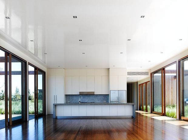 Bright, open-plan contemporary kitchen and living space with extensive floor-to-ceiling glazing, white flat-panel cabinetry, a dark stone backsplash and benchtop, polished dark timber flooring, and a high glossy white ceiling with recessed downlights. Timber-framed glass doors open toward landscaped outdoor areas.