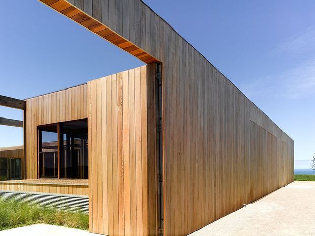 A contemporary coastal building exterior clad in vertical timber boards, with a strong angular roof overhang, recessed dark-framed glazing, and a pale paved walkway. The ocean is visible in the distance beyond the building, while low coastal planting borders the foreground.