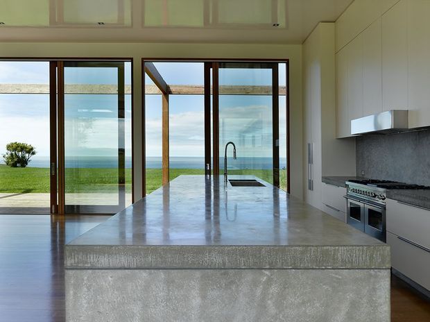 Contemporary open-plan kitchen with a large polished concrete island and integrated sink, facing expansive glazed doors with ocean views. White built-in cabinetry, a dark textured backsplash, stainless-steel appliances, timber-framed glazing, and a covered outdoor structure are visible.