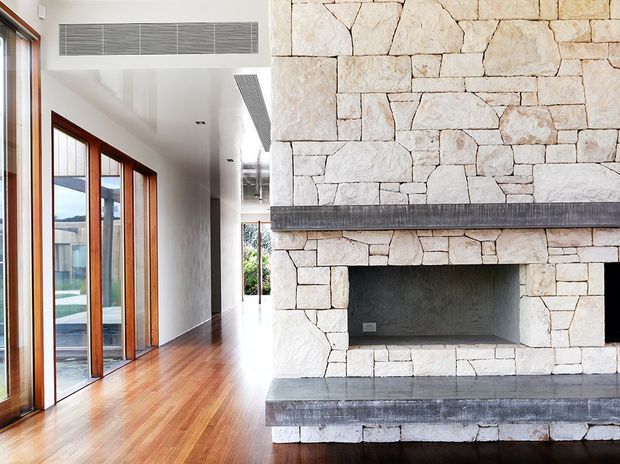 Bright contemporary interior featuring a large rustic split-stone fireplace with dark stone hearth and mantel, warm timber flooring, white walls, and full-height timber-framed glazing along a corridor-like living space.