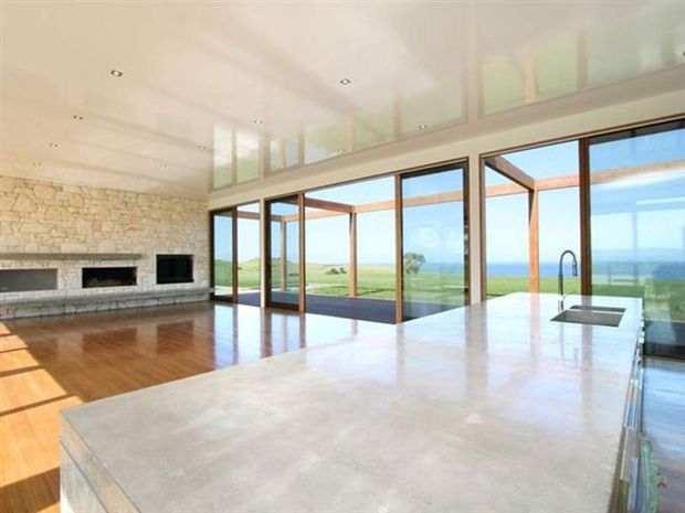Open-plan contemporary living and kitchen space with a large pale stone island in the foreground, warm timber flooring, a linear stone fireplace wall, and extensive floor-to-ceiling glazing. The glazing opens to a broad coastal landscape and ocean view, with clerestory windows above and exposed timber framing outside.