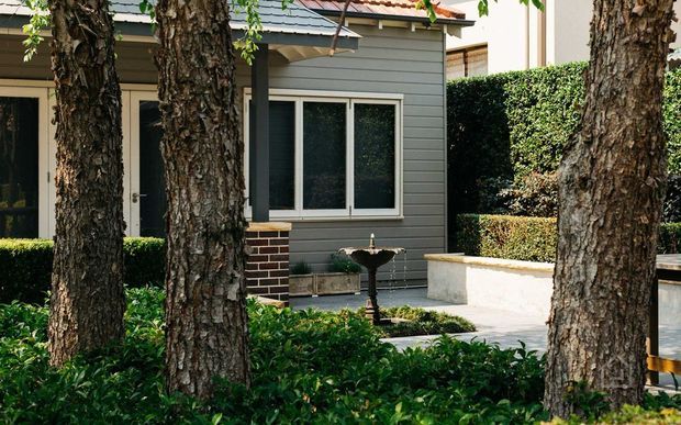 Exterior view of a compact residential building partly screened by mature trees and hedges. The facade combines dark grey horizontal weatherboards, cream-framed windows, and a lower section of dark brick, with a small fountain in a paved landscaped courtyard.