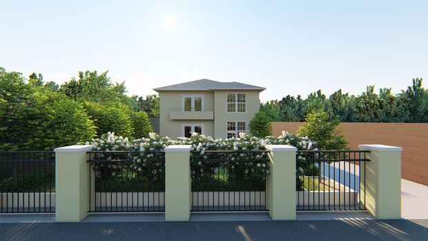 Exterior view of a two-storey residential house beyond a landscaped front boundary. The home has a simple hipped roof, light-rendered walls, vertically proportioned windows, and a balcony, with a black metal fence, rendered masonry posts, flowering planting, and a paved driveway in the foreground.