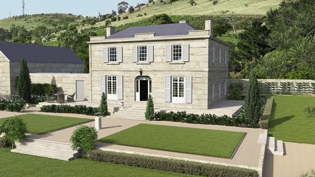 A formal two-storey stone residence set within a carefully landscaped hillside property. The symmetrical facade features pale ashlar stone walls, dark metal pitched roofing, white multi-pane shutters and windows, a central arched entrance, broad entry steps, clipped hedges, geometric lawns, and cypress-like planting. A matching outbuilding is visible to the left, with rolling green hills in the background.
