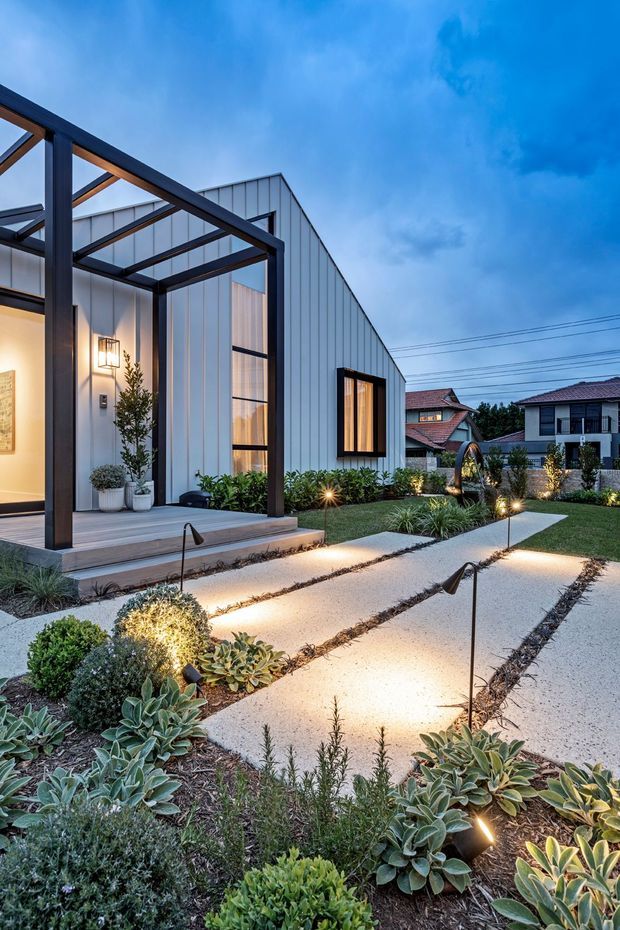 Dusk exterior view of a contemporary single-storey home with a light vertical-panel facade, dramatic dark steel-framed covered entry structure, warm interior lighting, and a landscaped front garden. Broad pale concrete stepping paths are separated by planted strips and illuminated with low garden lights. Nearby suburban homes and overhead utility wires are visible in the background.