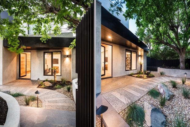 A contemporary single-storey home exterior shown in a split view, with dark horizontal cladding and trim, textured light rendered walls, black-framed glazing, and warm interior light visible through the windows and door. A landscaped front courtyard uses pale gravel, large stepping slabs, boulders, ornamental grasses, and mature trees.