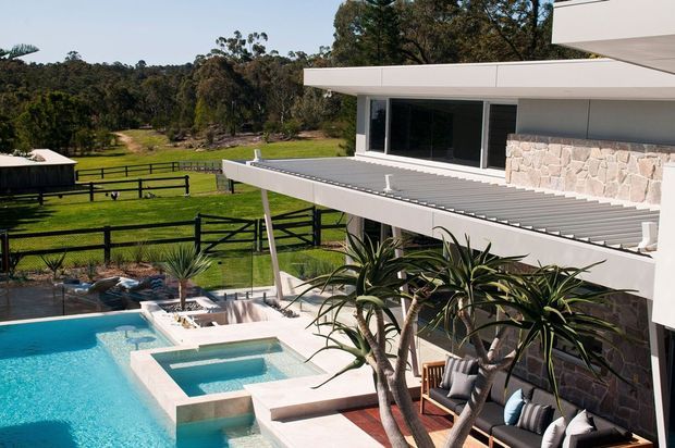 Contemporary multi-level residence overlooking a landscaped rural property, with extensive white rendered forms, broad glazing, a stone feature wall, covered outdoor entertaining area, and a swimming pool with integrated spa. The image is viewed from outside and primarily showcases the building facade, roof forms, pool terrace, and surrounding grounds.