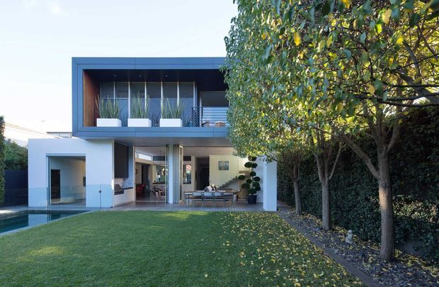 Contemporary two-storey residence viewed from a landscaped backyard, featuring a crisp white rendered ground floor, dark charcoal-blue rectangular upper-level frame, extensive glazing, balcony planters, and a covered outdoor entertaining area. A lawn, swimming pool edge, and mature trees frame the home.