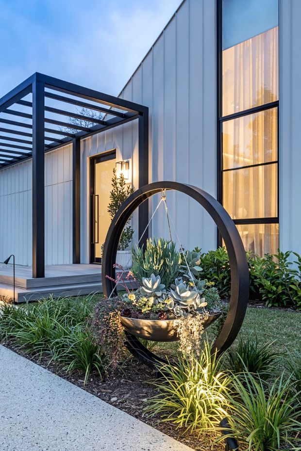 Contemporary residential entry viewed from the landscaped front walkway, featuring a crisp white vertically clad facade, tall black-framed glazing, a covered entrance canopy with dark structural framing, and a sculptural circular planter filled with succulents and ornamental grasses.
