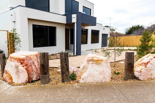 Exterior view of a contemporary two-storey residence with white rendered walls, dark charcoal window frames and projecting upper-level forms. The front landscape features large natural stone boulders, timber posts, gravel and sparse planting, with a timber boundary fence behind.