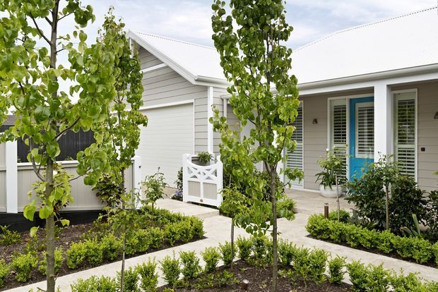 Street-facing residential exterior with light grey horizontal weatherboard cladding, a white pitched roof, white-framed windows with shutters, and a vivid blue entry door. A paved garden path crosses through a formal landscaped front garden with young trees, clipped hedges, and low planting. The building has a clean, traditional coastal character.